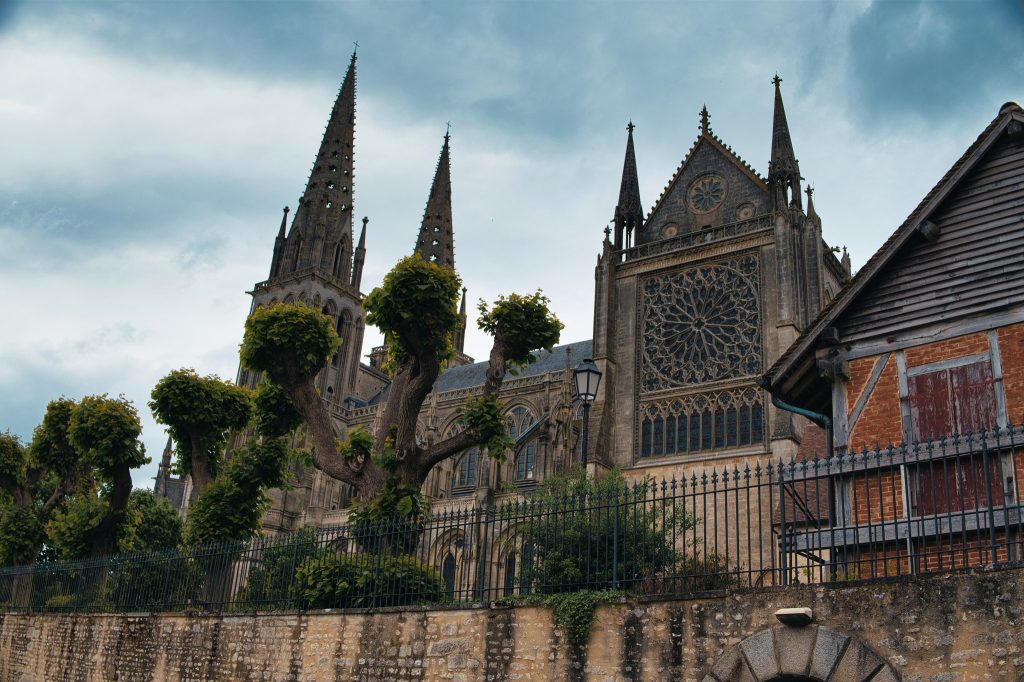 Flèches et rose du transept sud de la cathédrale Notre-Dame de Sées derrière des arbres taillés et une grange, dans l'Orne
