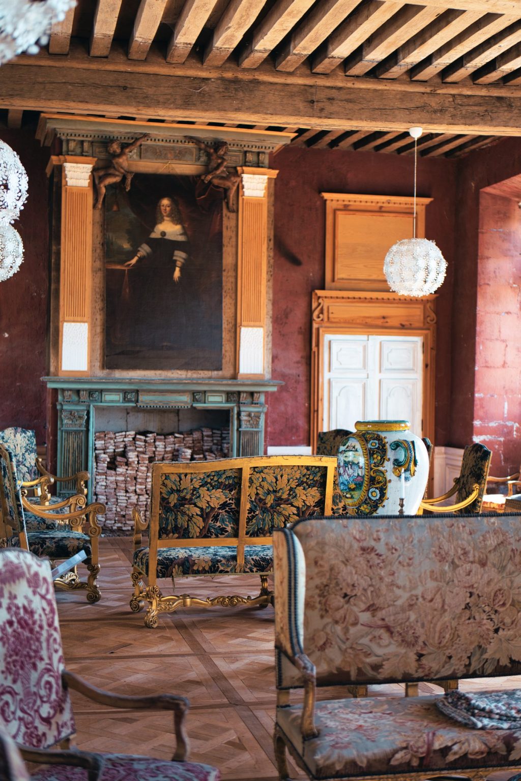 Salon de château aux poutres apparentes, cheminée surmontée d'un portrait ancien, fauteuils tapissés et vase de faïence