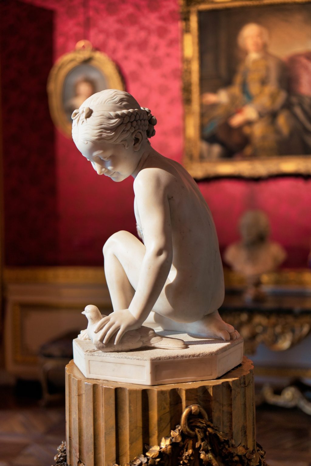 Sculpture en marbre blanc d'une jeune fille accroupie tenant une colombe, devant un mur tendu de damas rouge et des portraits