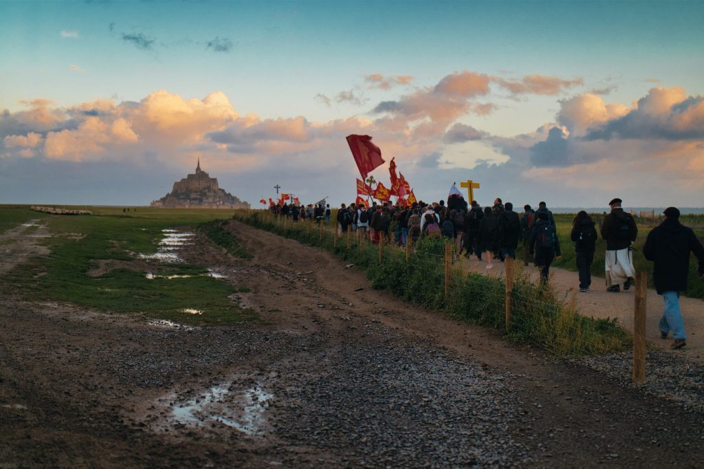 Colonne de pèlerins portant drapeaux rouges et croix sur un chemin des herbus, face au Mont-Saint-Michel au lever du jour
